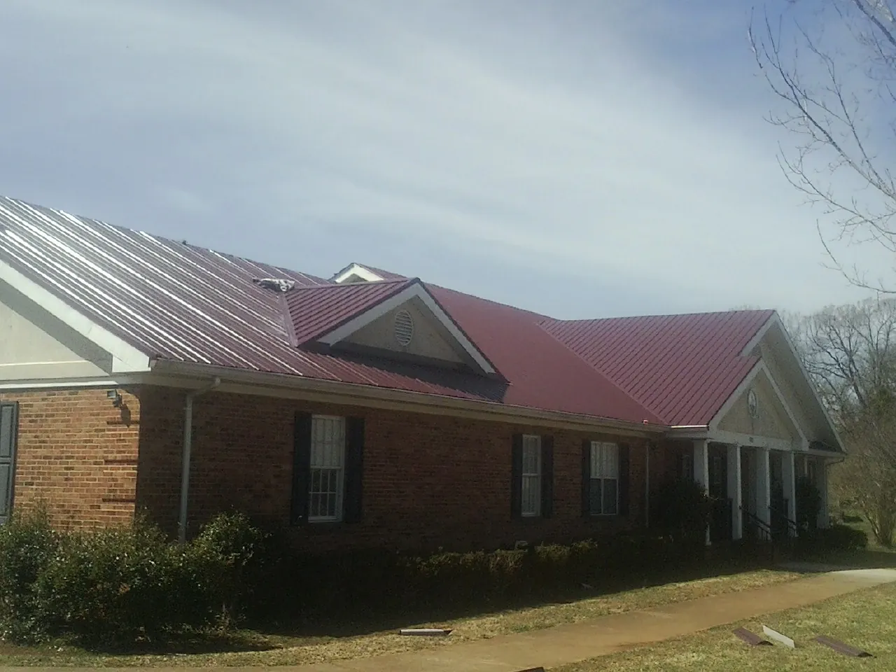 Red metal roof with gabled entries on a long brick building, from a Hail Damage Roof Repair job in Trafford