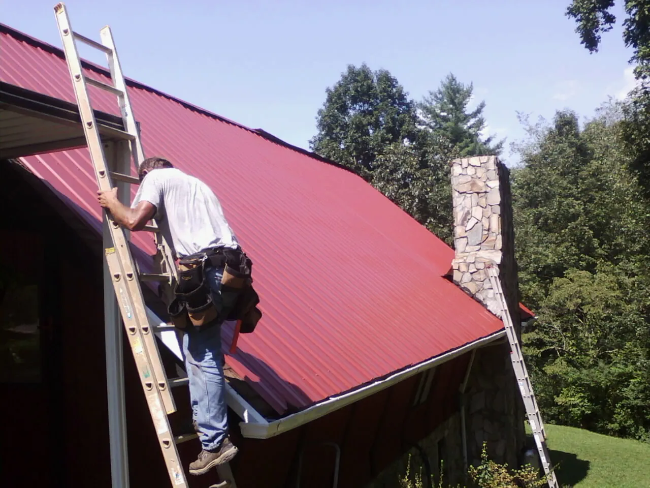 Roofer on a ladder beside a red metal roof and stone chimney — Soffit & Fascia Repair by Roofing Brothers in Trafford